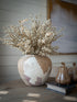 A rustic ceramic vase with a distressed beige, white, and muted mauve glaze, filled with faux soft cream heather spray, displayed on a wooden table next to decorative pumpkins and a book.