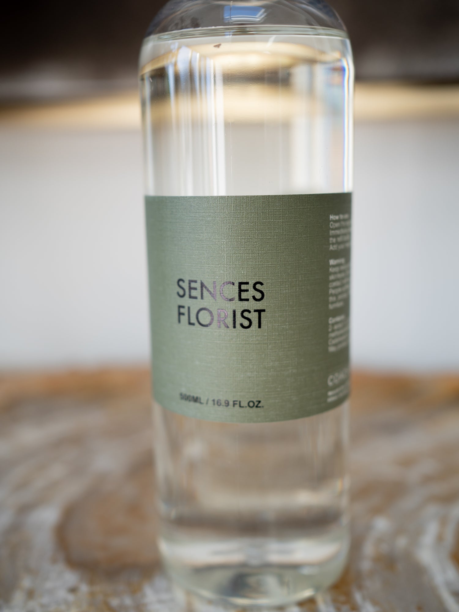 A clear glass bottle of Sences Florist refill bottle with a muted sage green label, resting on a rustic wooden surface with a softly blurred background.