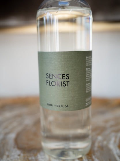 A clear glass bottle of Sences Florist refill bottle with a muted sage green label, resting on a rustic wooden surface with a softly blurred background.