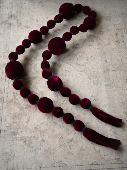 A long beaded garland made of deep burgundy velvet beads and tassels, laid out on a textured beige surface.