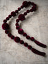 A long beaded garland made of deep burgundy velvet beads and tassels, laid out on a textured beige surface.
