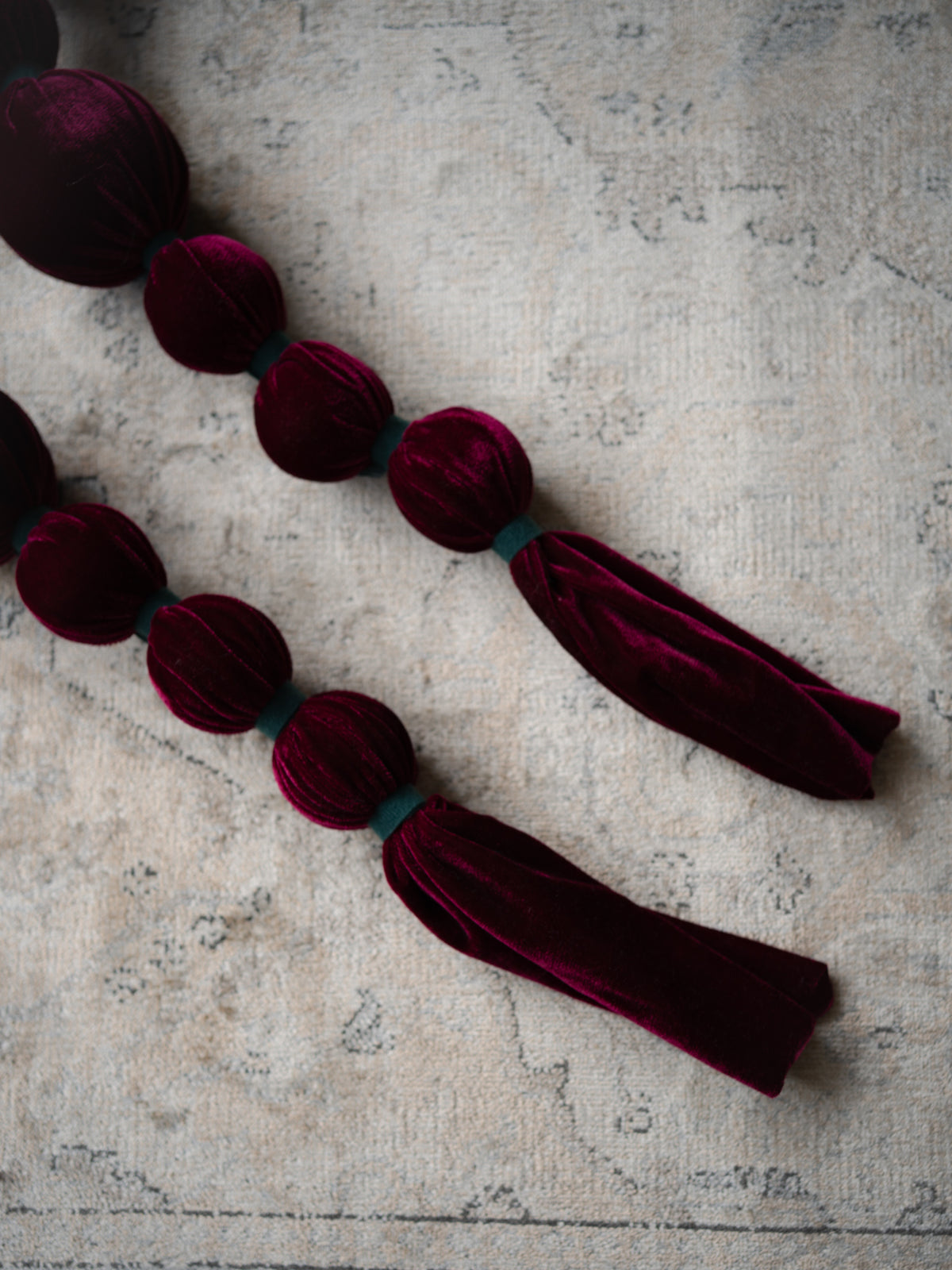 Deep burgundy velvet tassel garland with knotted bead details, laid diagonally on a textured light gray stone surface.