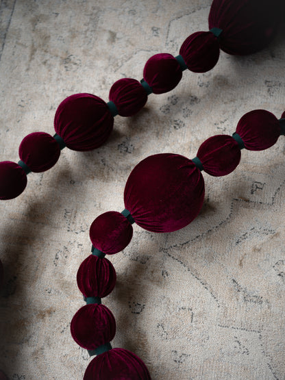 A close-up of a dark burgundy velvet garland with large round beads and small teal ribbon accents, laid diagonally across a textured beige surface.