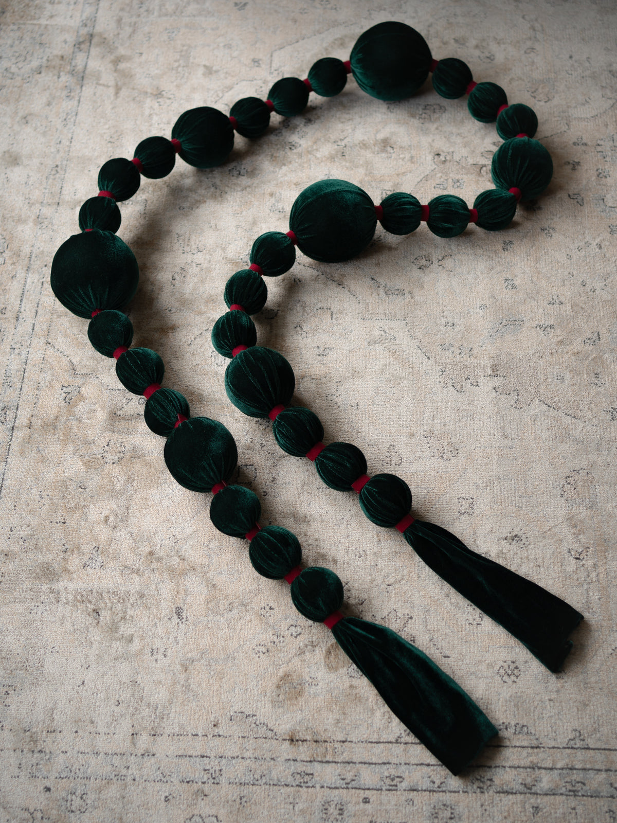 A dark green velvet beaded garland with alternating large and small round beads, accented by thin red threads between the beads, finished with two long dark green velvet tassels, laid out on a textured beige surface.