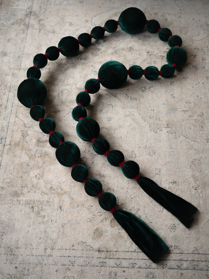 A dark green velvet beaded garland with alternating large and small round beads, accented by thin red threads between the beads, finished with two long dark green velvet tassels, laid out on a textured beige surface.