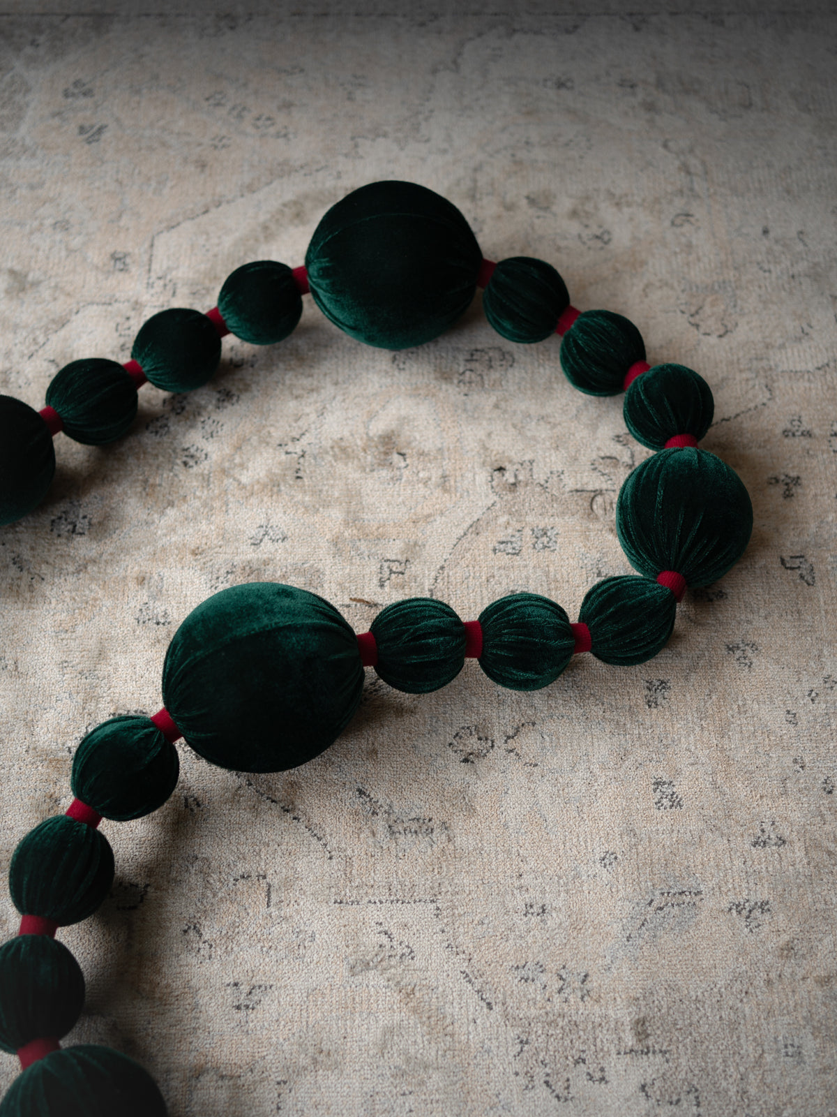 A beaded garland featuring large dark green velvet beads separated by small red ribbon ties, laid out on a textured beige fabric background.