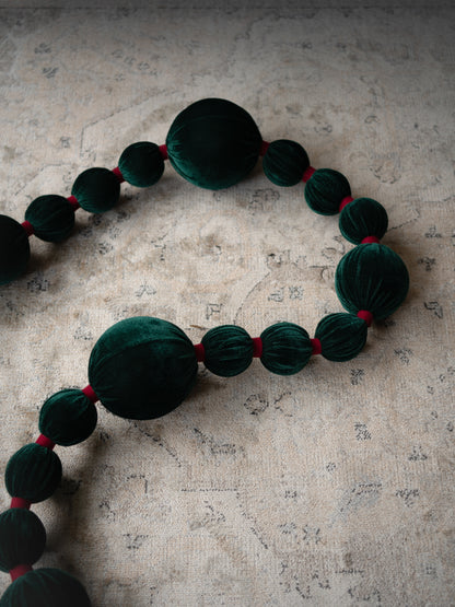 A beaded garland featuring large dark green velvet beads separated by small red ribbon ties, laid out on a textured beige fabric background.