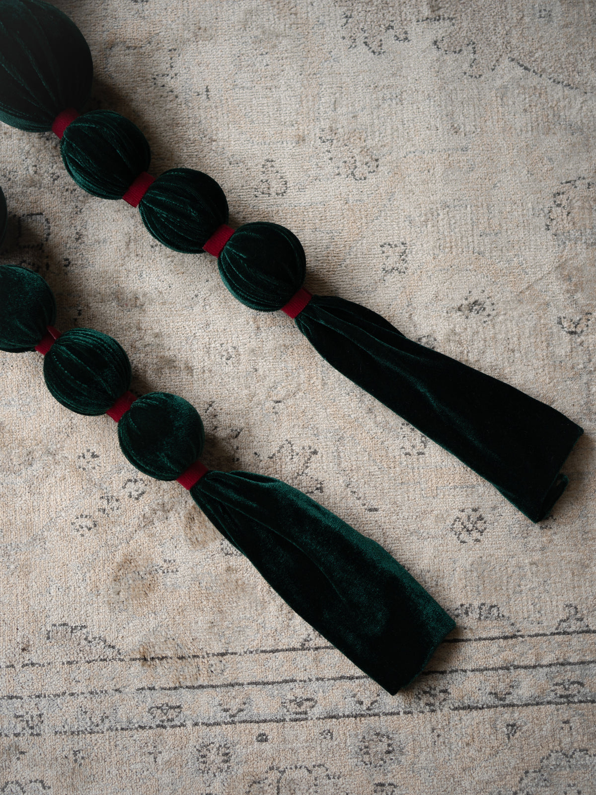 Dark green velvet tassel garland with round bead-like segments separated by thin red bands, laid diagonally on a textured beige fabric background.