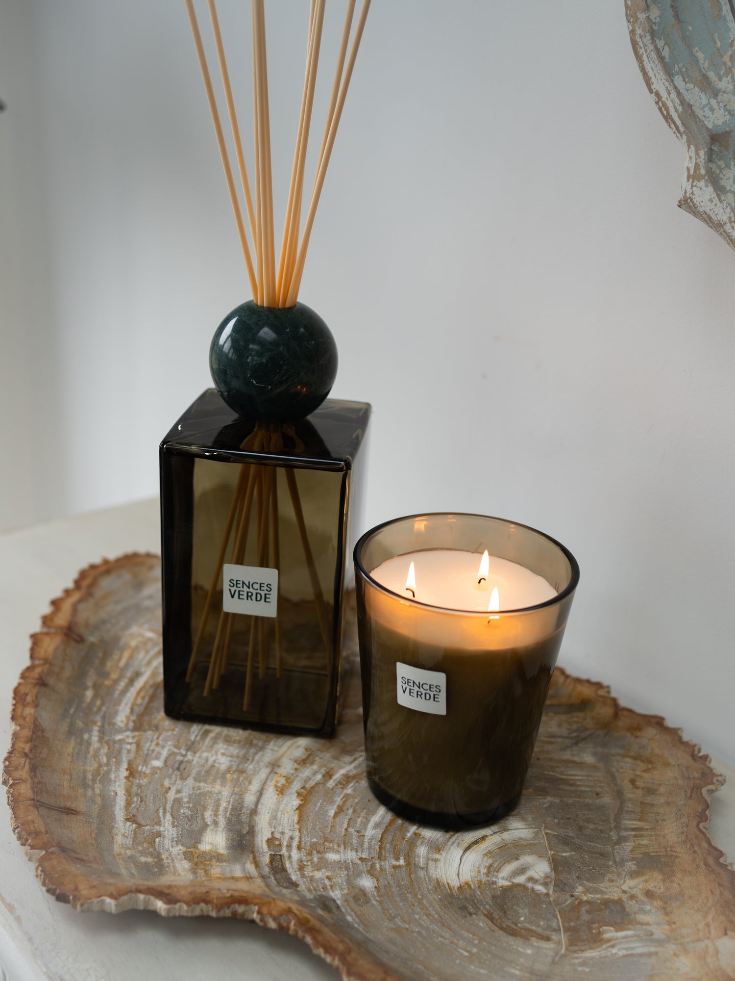 A lit green glass candle in a matching amber glass holder sits beside a dark amber reed diffuser bottle on a rustic fossiled wood slice tray against a light gray wall.