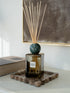 A reed diffuser bottle with a dark green marble sphere and natural reeds, placed on a stone tray in a styled interior setting.