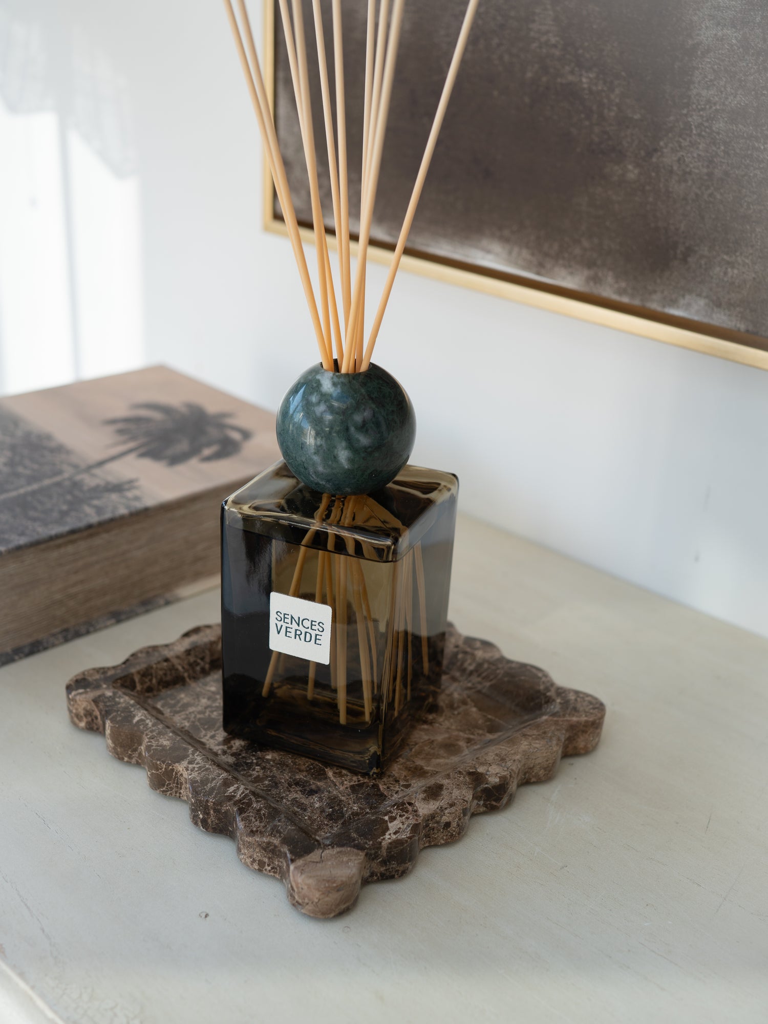 A reed diffuser with a dark green marble sphere and natural reeds, set in a square dark glass bottle on a rustic stone coaster. The bottle has a white label reading &