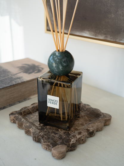 A reed diffuser bottle with a dark GREEN glass body, a round green marble sphere, and natural reeds, resting on a decorative stone tray in a styled interior setting.