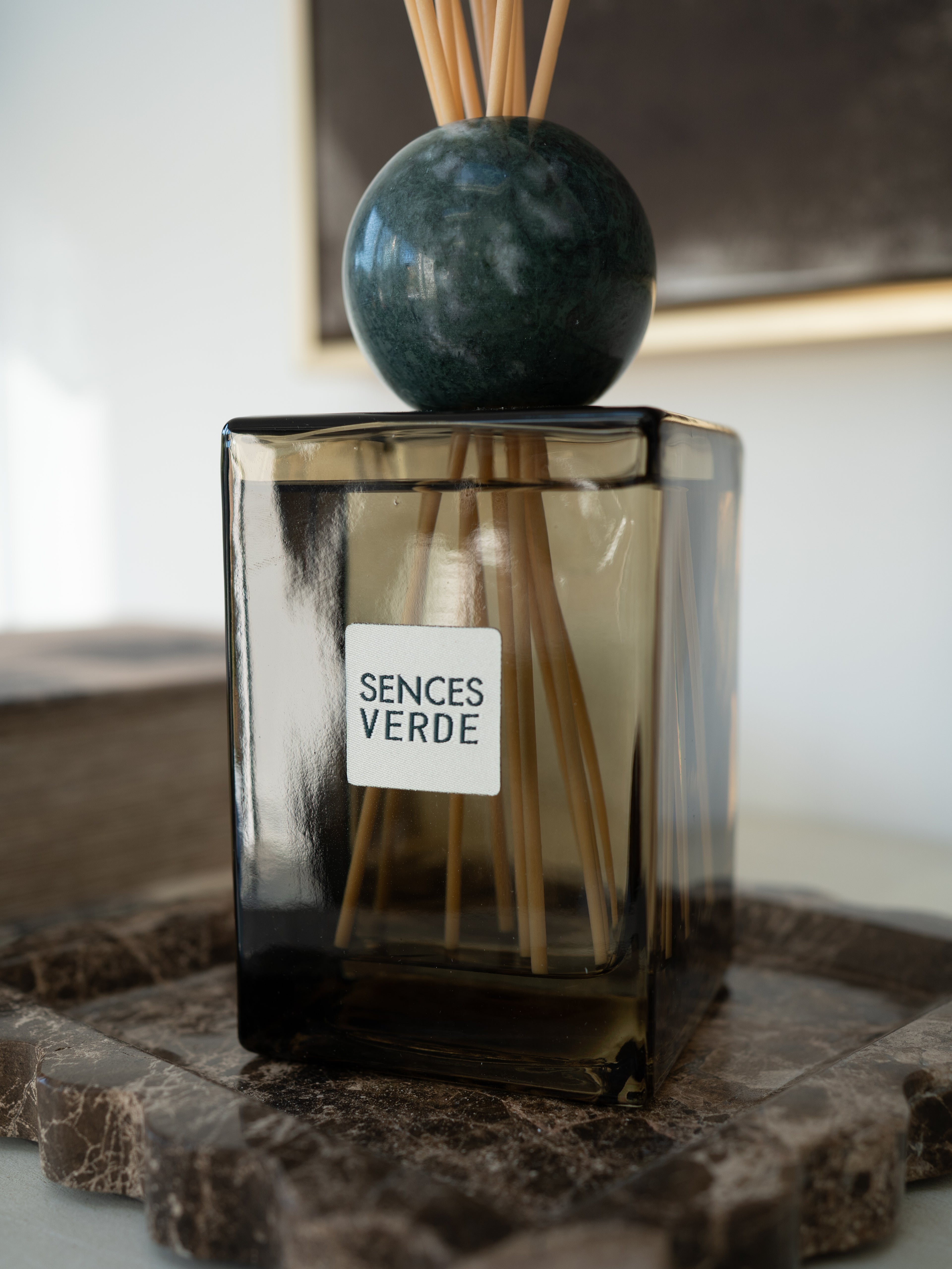 A Sences Verde reed diffuser with a square dark green glass bottle, a round dark green marble top, and natural wood reeds, resting on a stone tray.