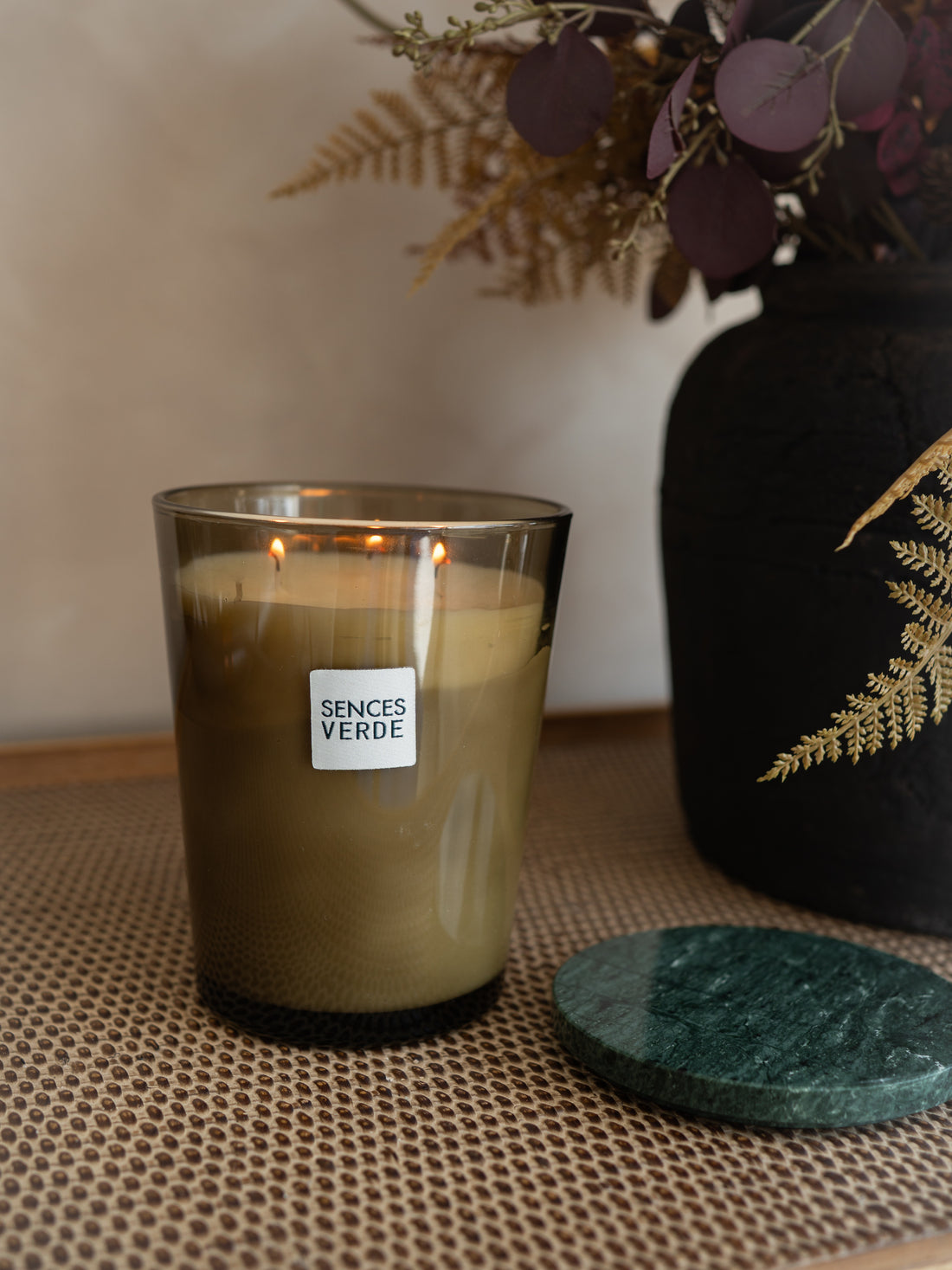 A lit Sences Verde candle in a clear glass jar sits on a woven beige mat, with a dark green round coaster beside it and a black decorative vase with dried foliage in the background.