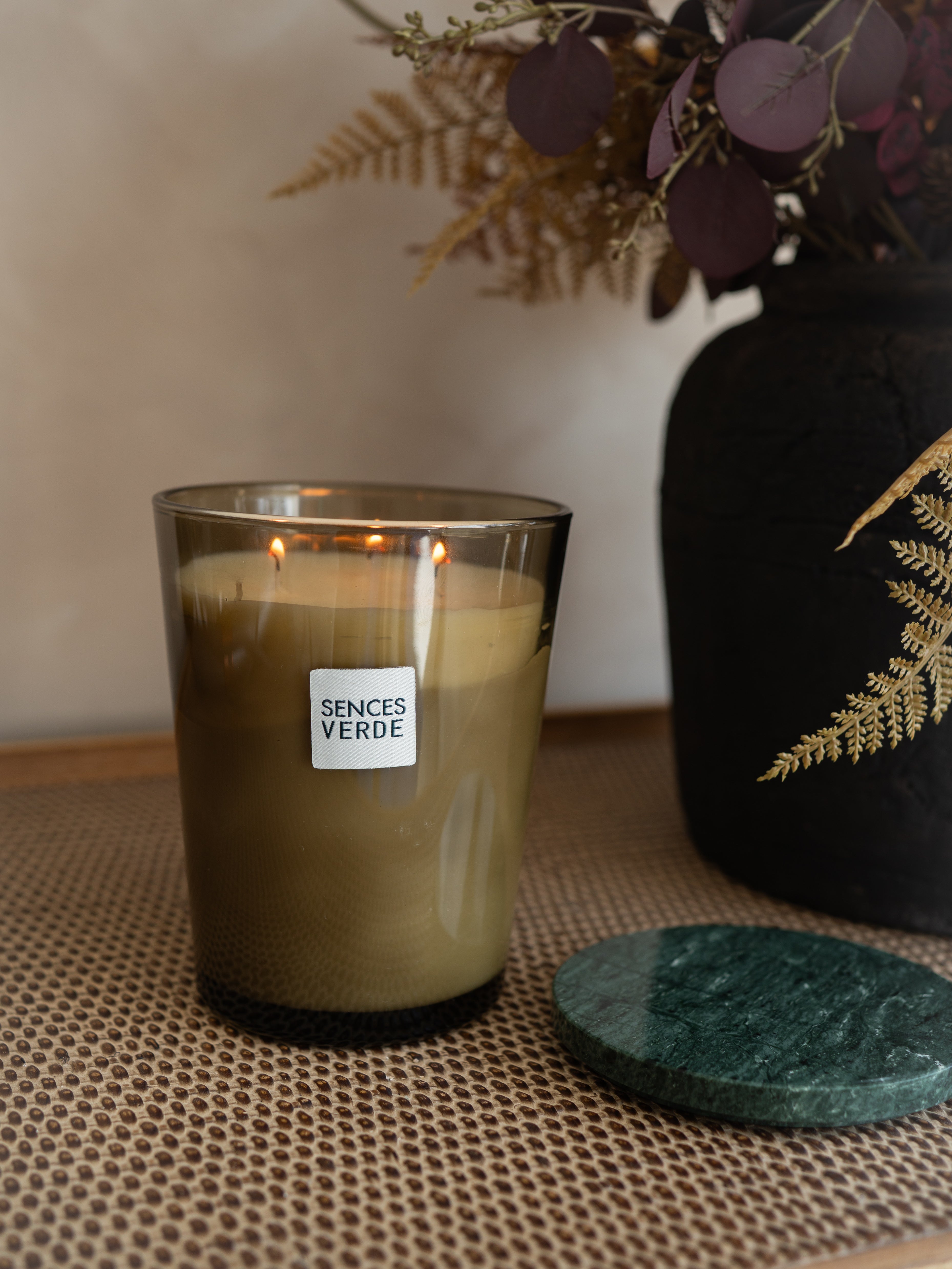 A lit Sences Verde candle in a clear glass jar sits on a woven beige mat, with a dark green round coaster beside it and a black decorative vase with dried foliage in the background.