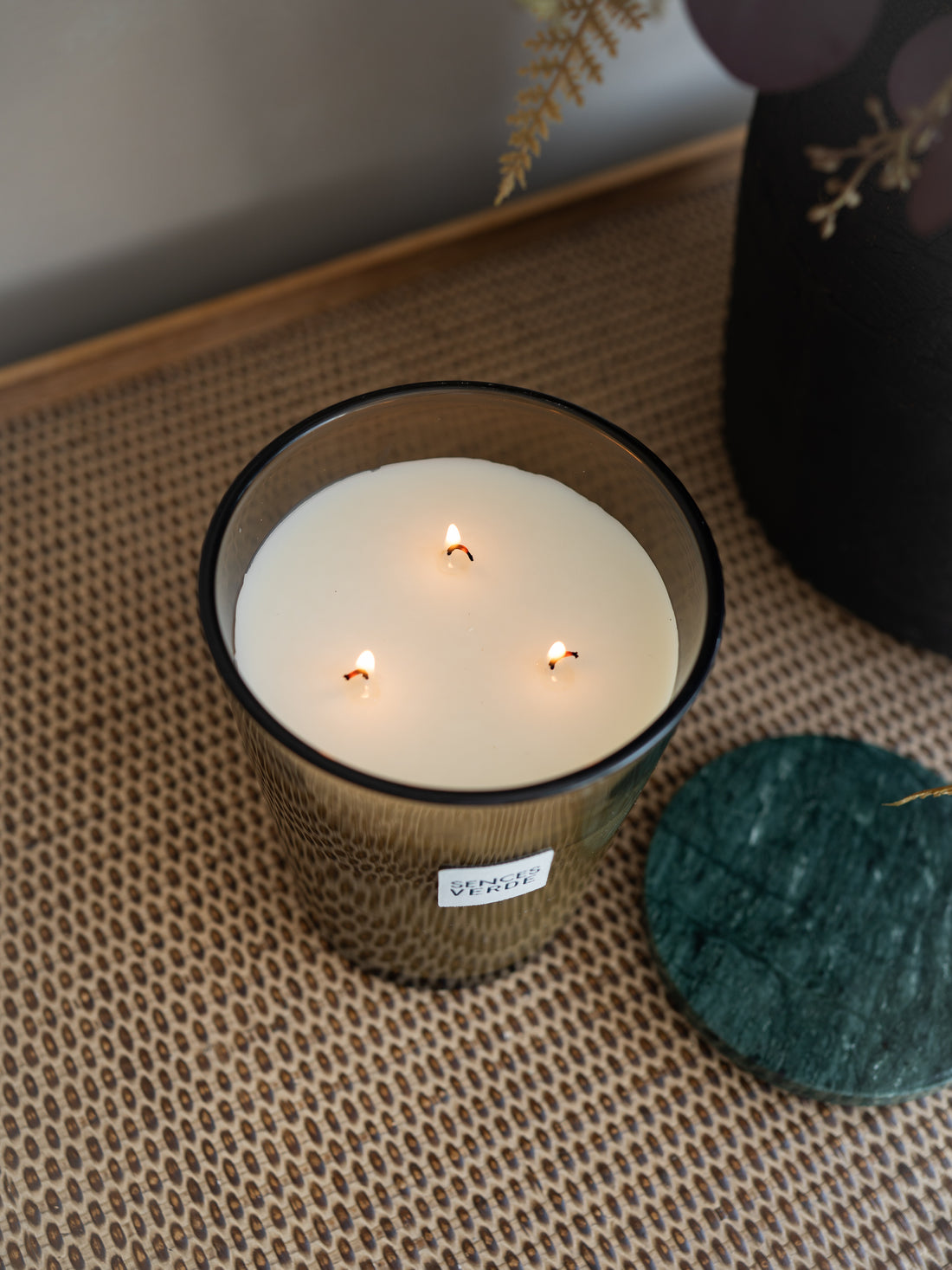 A lit three-wick candle in a dark amber glass jar sits on a woven beige mat, with a dark green stone coaster beside it and a blurred black vase in the background.