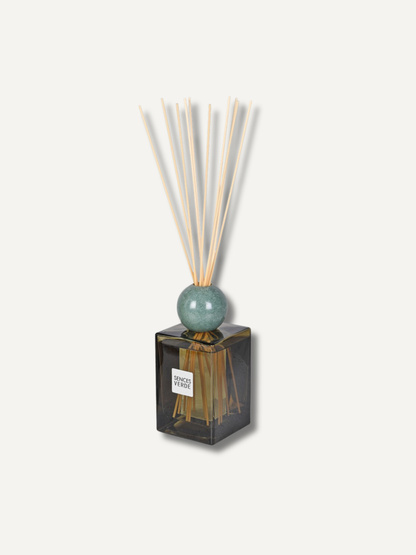 A reed diffuser with a square dark green glass bottle, a round pale green top, and natural reed sticks, shown on a light beige background.
