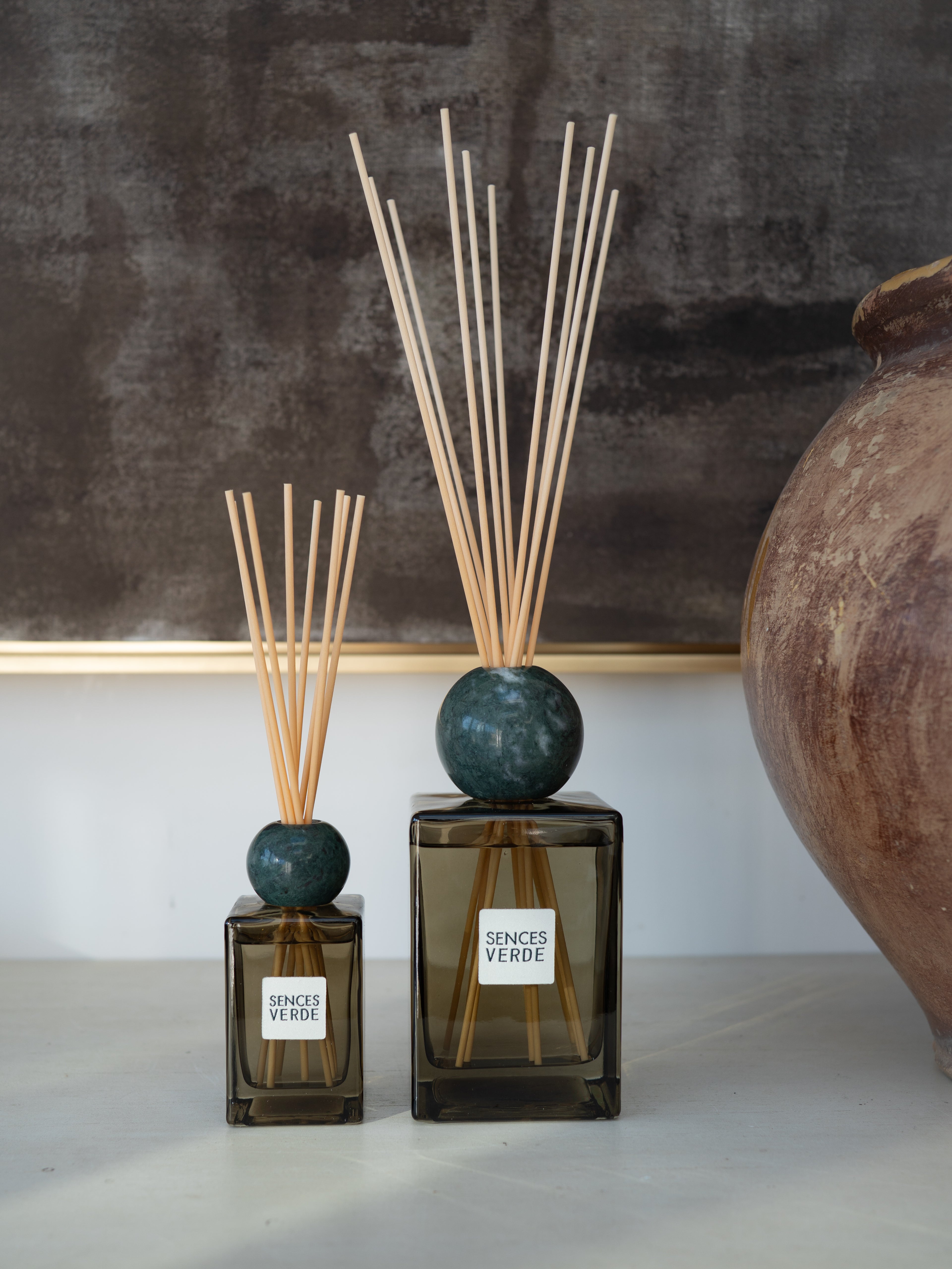 Two reed diffusers with dark green glass bases, round dark teal stoppers, and natural reed sticks, displayed on a light surface against a dark textured wall with a large ceramic pot partially visible on the right.