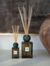 Two reed diffusers with dark green glass bases, round dark teal stoppers, and natural reed sticks, displayed on a light surface against a dark textured wall with a large ceramic pot partially visible on the right.
