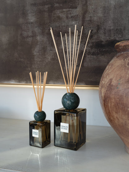 Two reed diffusers with dark green marble spheres and natural wood reeds, displayed on a light gray surface against a dark textured wall with a terracotta vase on the right.