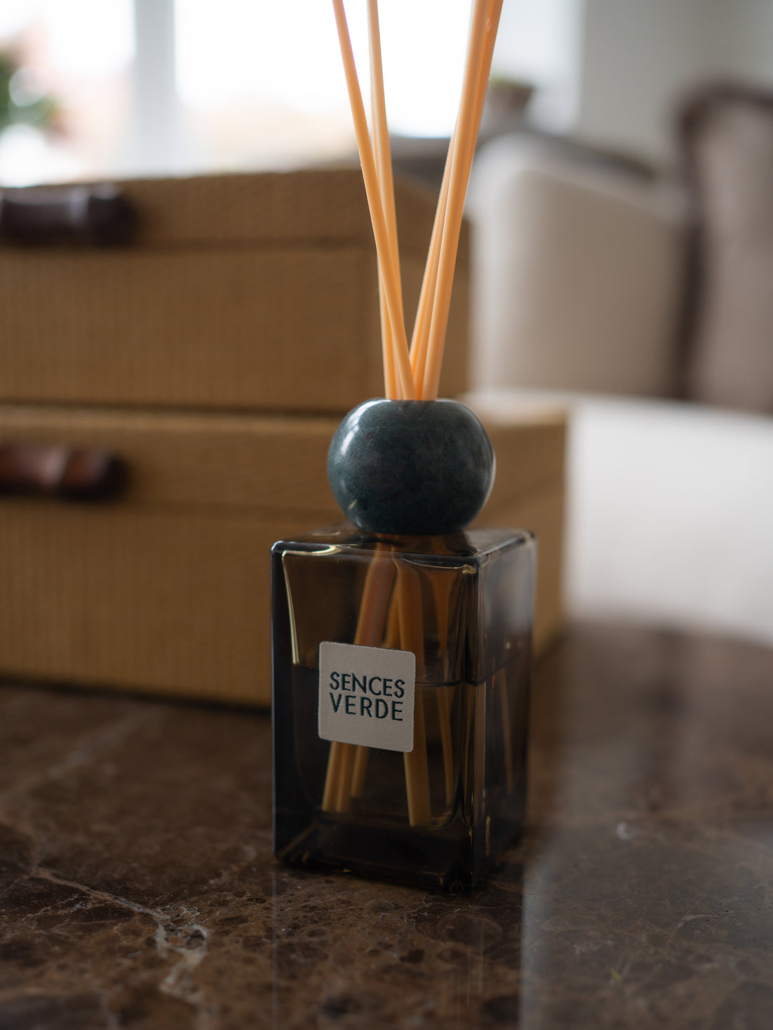 A reed diffuser bottle with a dark glass container, natural reeds, and a round dark blue-green marble stopper, labeled &