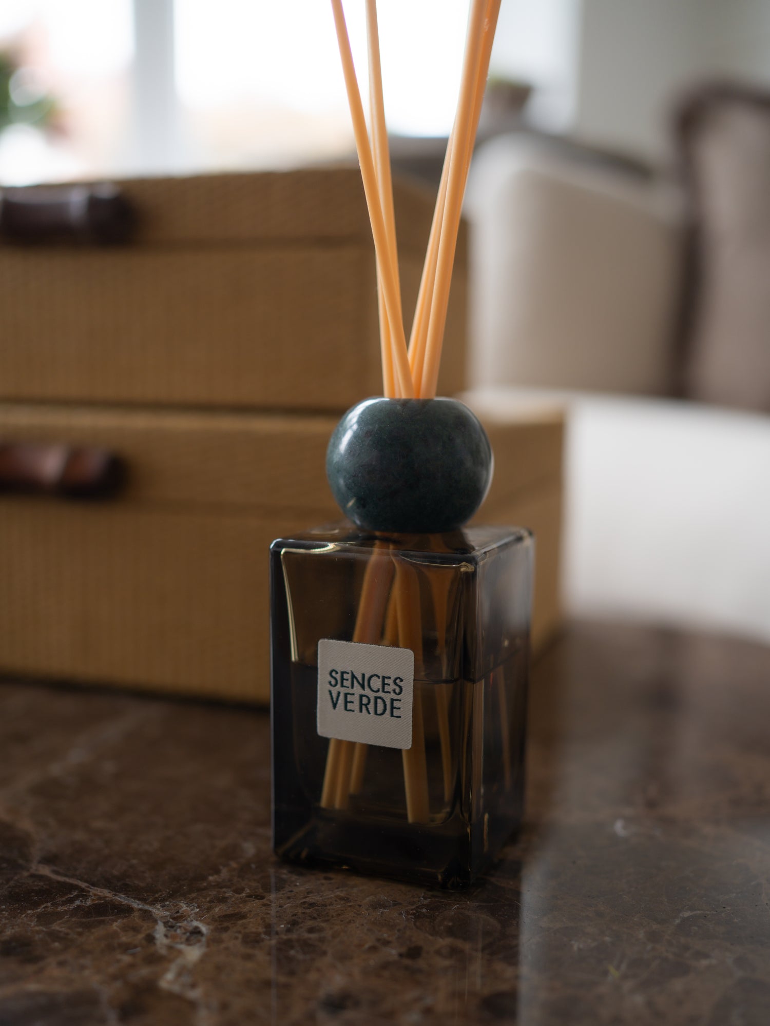 A reed diffuser bottle with a dark glass container, natural reeds, and a round dark blue-green marble stopper, labeled &