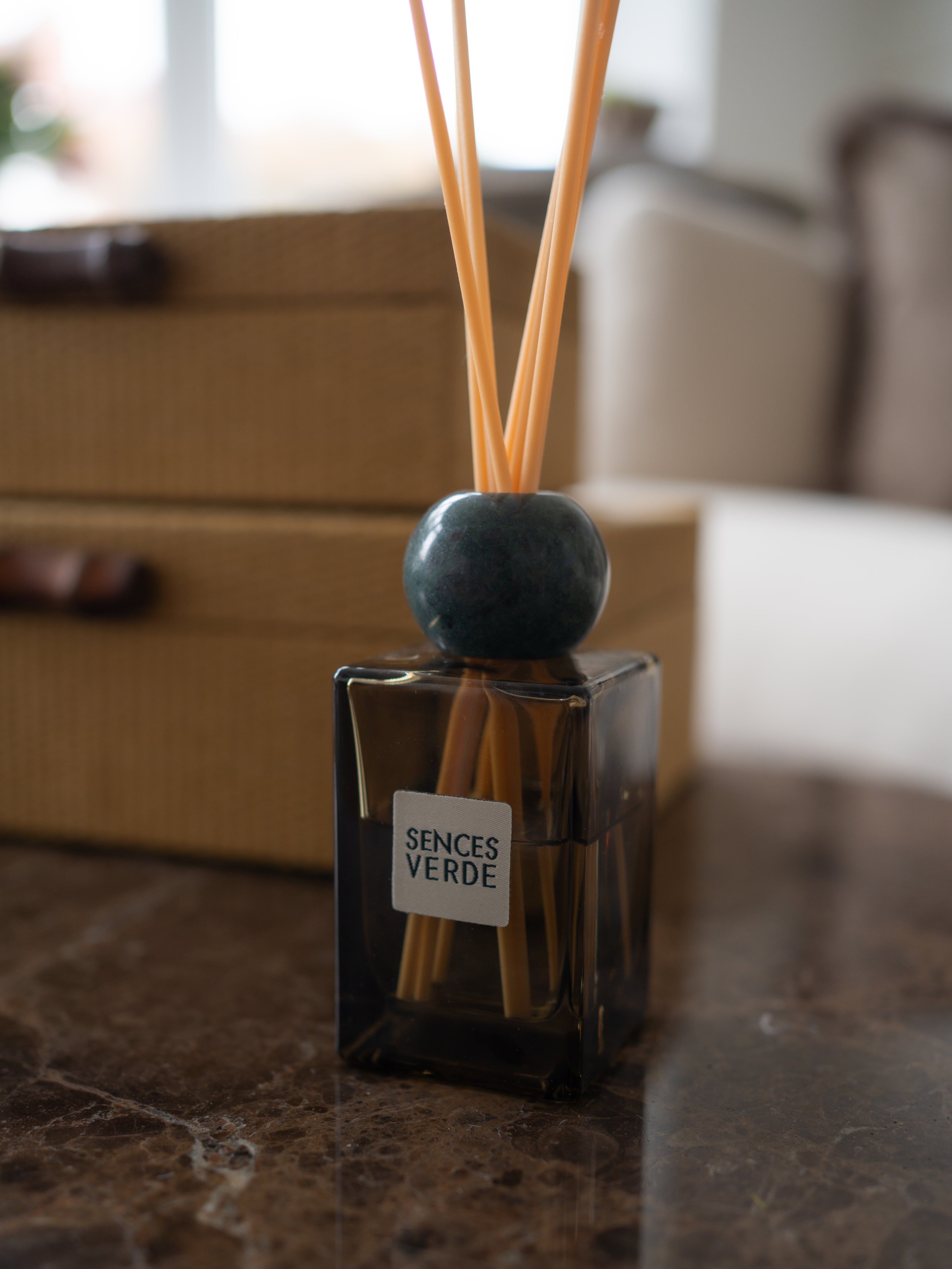 A reed diffuser bottle with a dark glass container, natural reeds, and a round dark blue-green marble stopper, labeled &