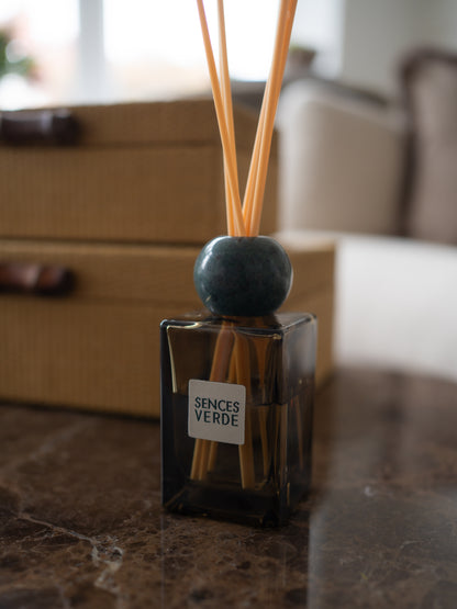 A reed diffuser bottle with a dark glass container, natural reeds, and a round dark blue-green marble stopper, labeled &