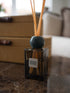 A reed diffuser bottle with a dark glass container, natural reeds, and a round dark blue-green marble stopper, labeled &