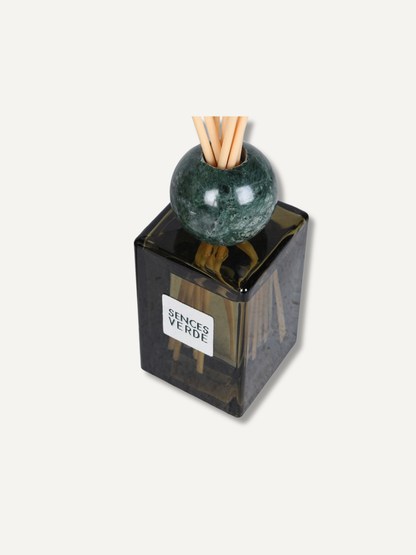 A reed diffuser bottle with a dark translucent square glass body, a green marble sphere, and natural reed sticks, with a white label reading &