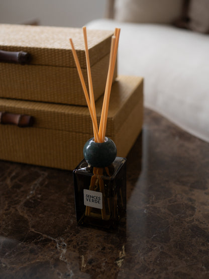 A reed diffuser bottle with light wood reeds and a dark round top, placed on a dark marble surface in front of woven storage boxes. The bottle label reads &