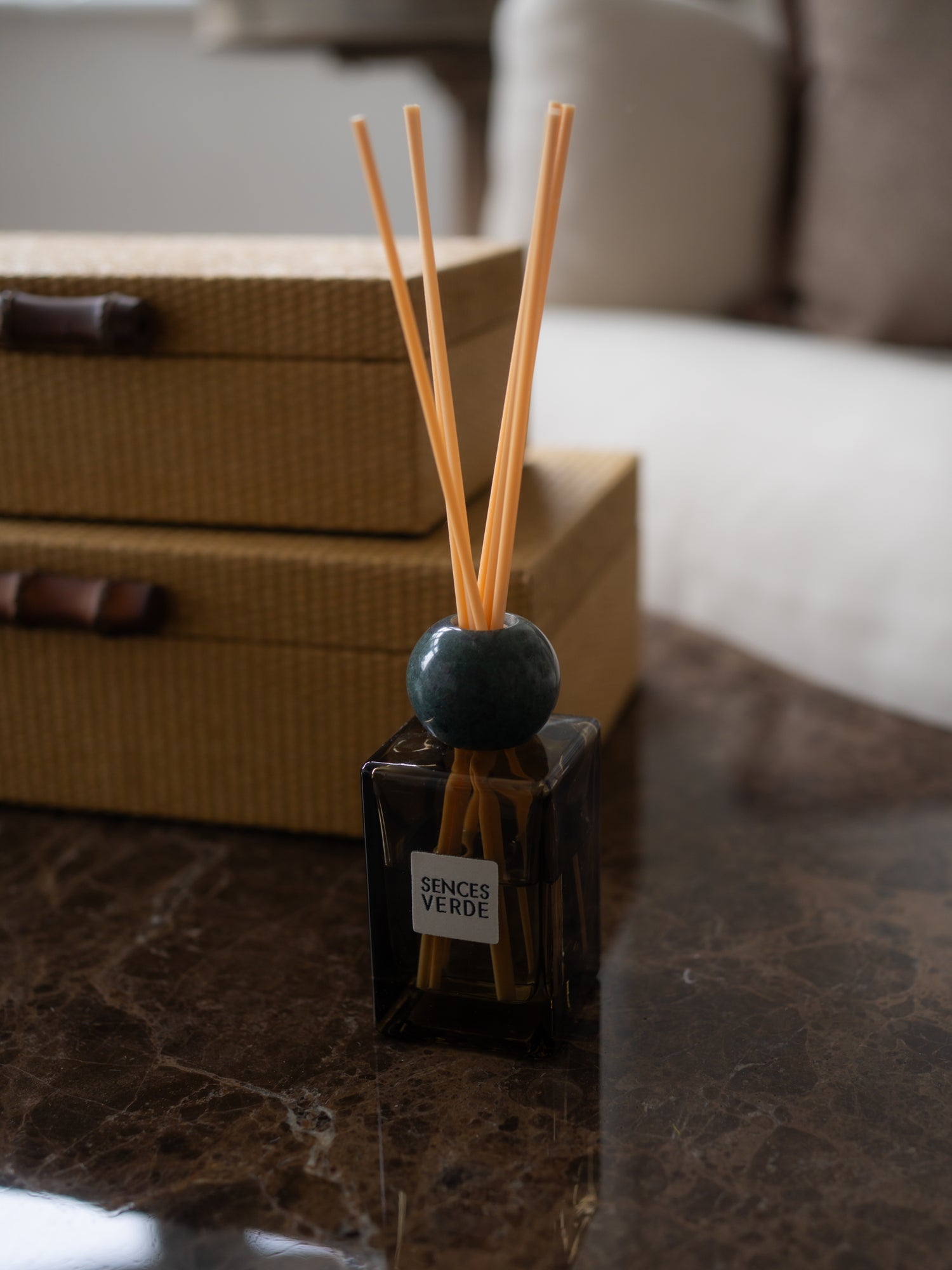 A reed diffuser bottle with natural reed sticks, a dark green spherical stopper, and a white label reading &