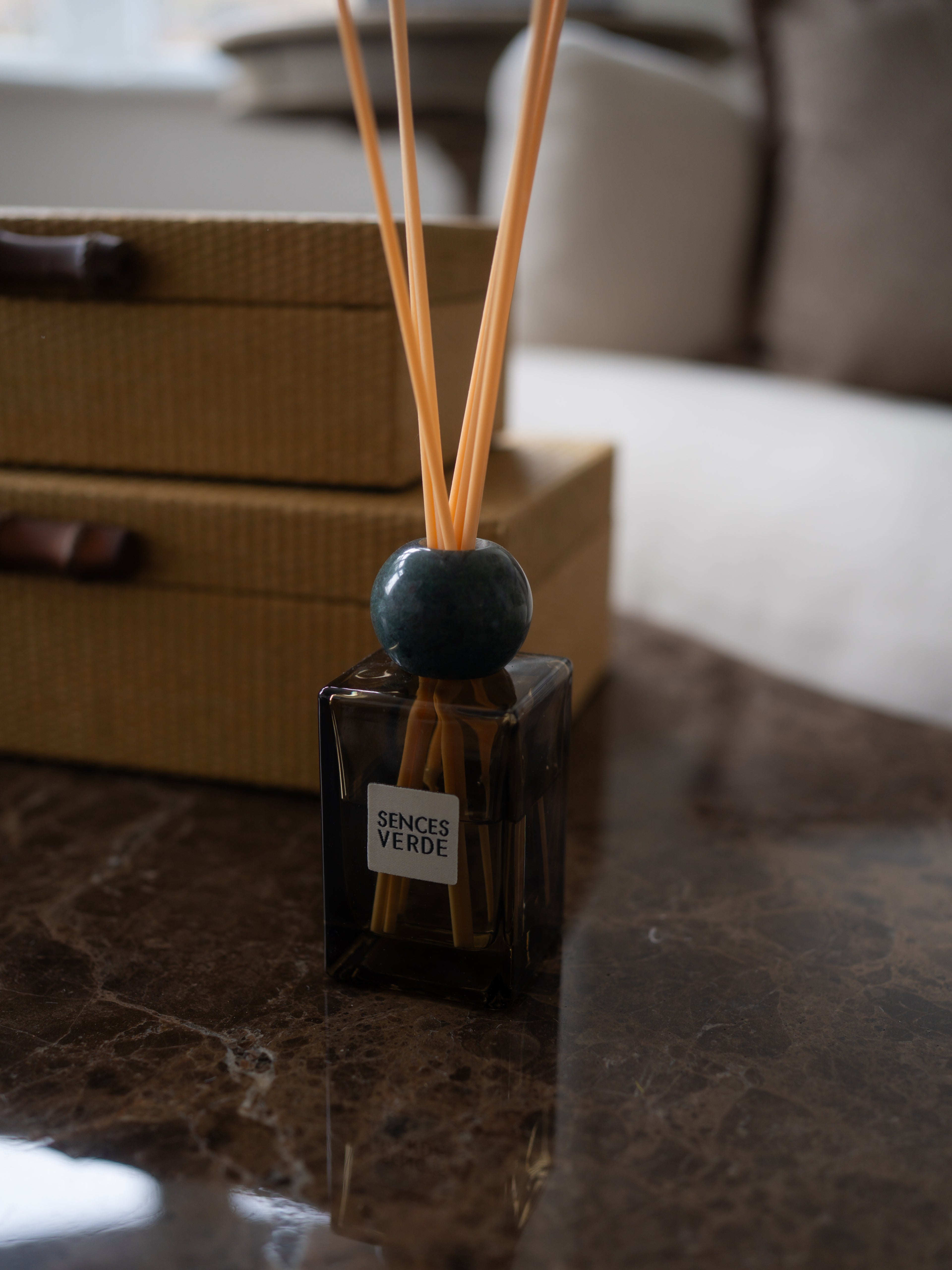 A reed diffuser bottle with a dark amber glass body, a round dark teal marble stopper, and natural reed sticks, placed on a polished dark marble surface with a woven box in the blurred background. The label reads &