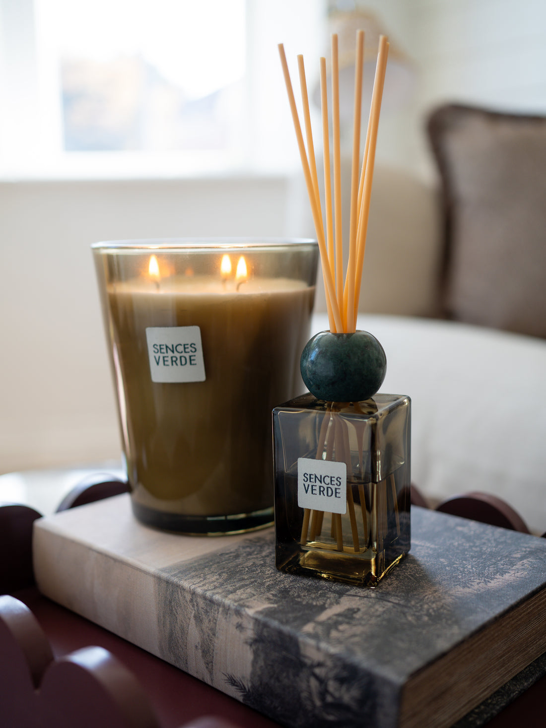 A lit candle and a reed diffuser set on a marble-patterned book in a softly blurred indoor setting. Both products have white labels reading &