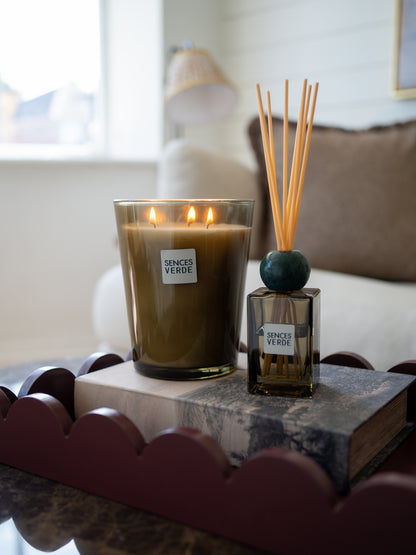 A lit scented candle in a clear glass jar and a matching reed diffuser bottle labeled &