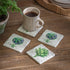 Set of four decorative coasters with plant designs on a wooden surface with a mug of coffee.