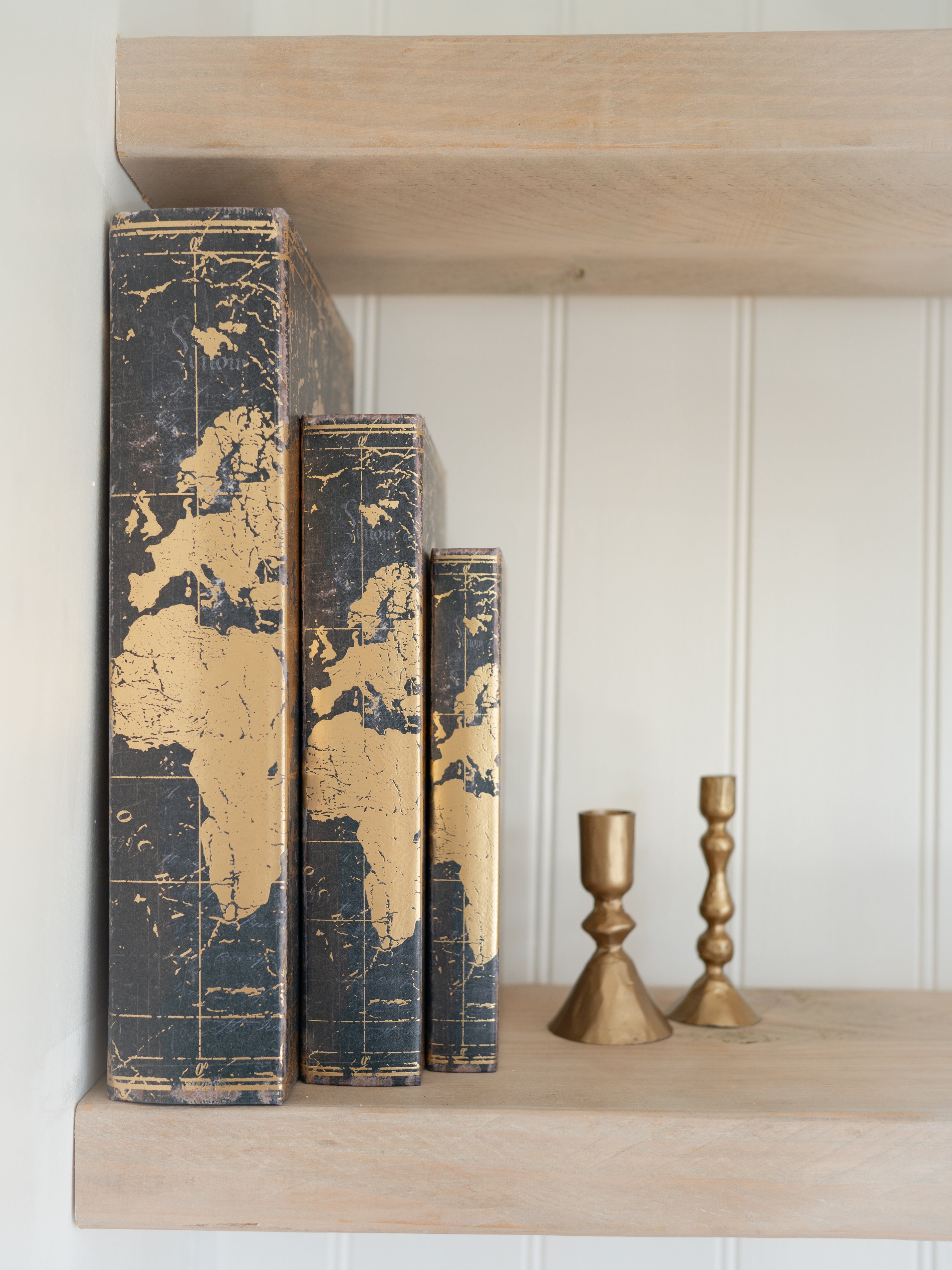 Decorative storage books with world map design on a wooden shelf with gold candle holders.