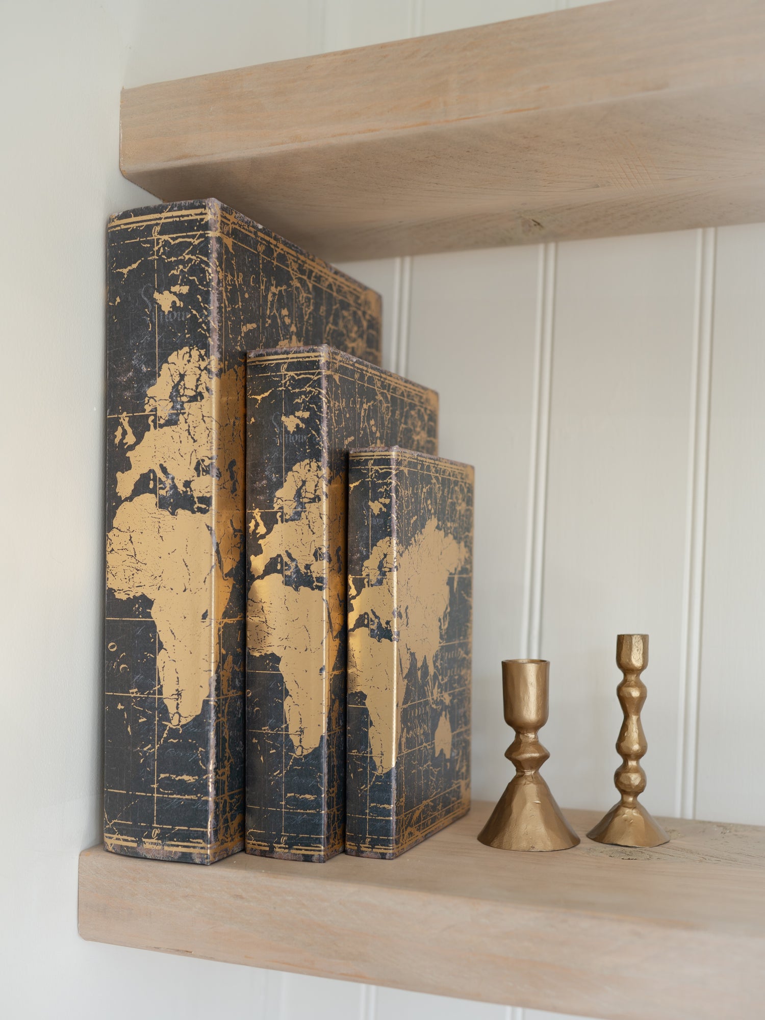 Three world map storage books on a wooden shelf with two gold candle holders.