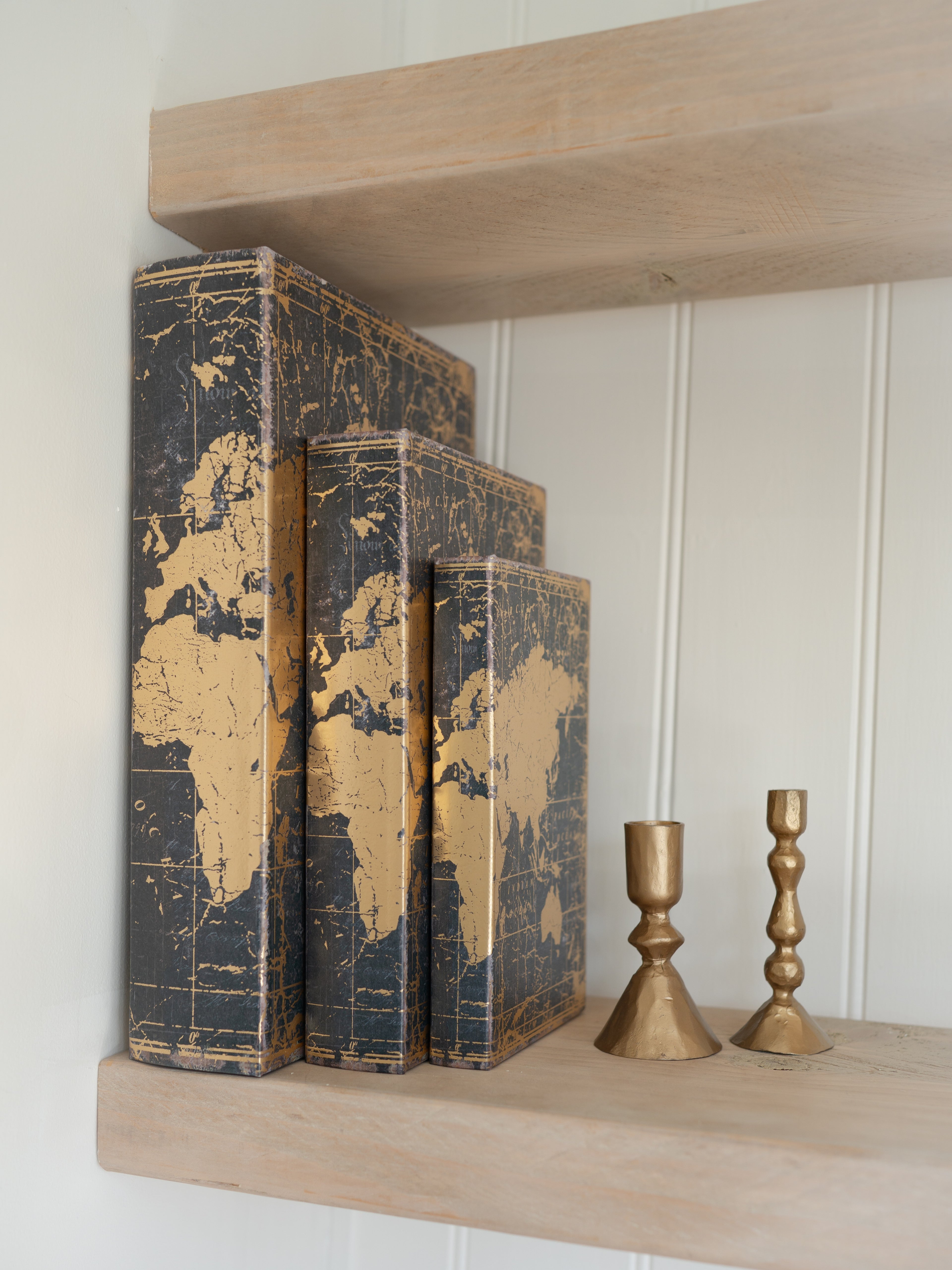 Three world map storage books on a wooden shelf with two gold candle holders.