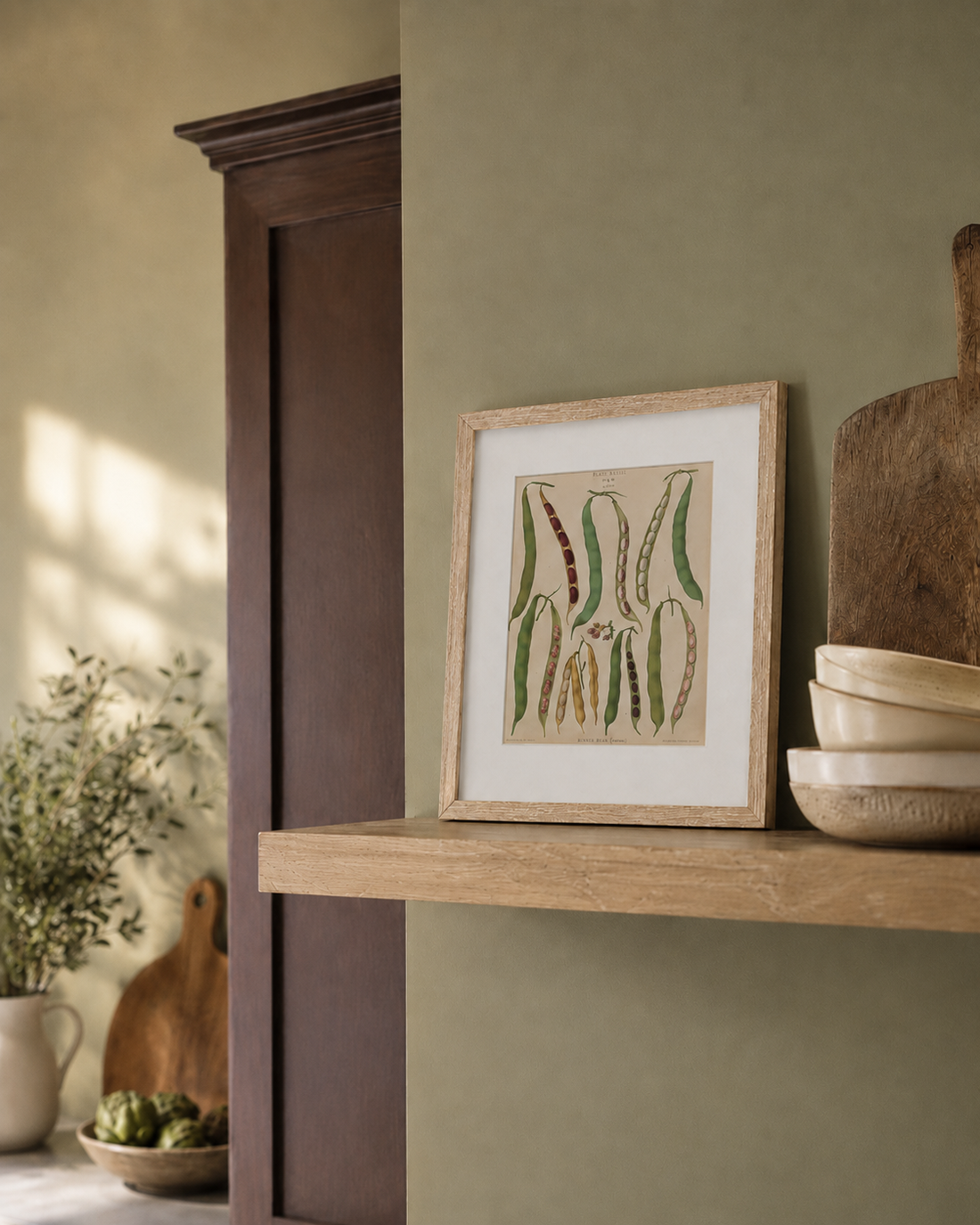 A framed botanical art print featuring green and brown beans on a beige background, styled on a light wood floating shelf next to stacked ceramic bowls and a wooden cutting board against a sage green wall.