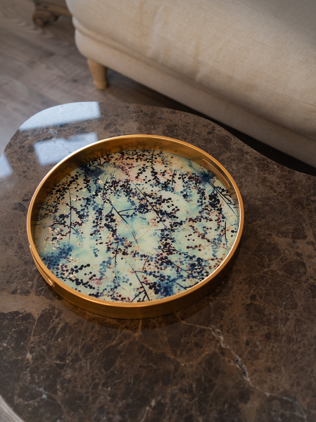 A round decorative tray with a gold-toned rim and a blue, cream, and black blossom pattern, resting on a dark marble coffee table in a living room setting.