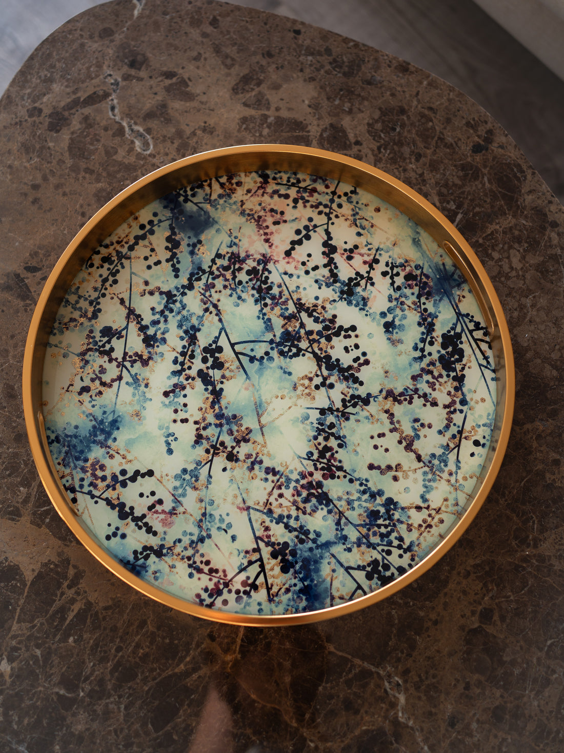 A round decorative tray with a gold-toned rim and a blossom pattern of blue, cream, and black, placed on a dark brown marble surface.