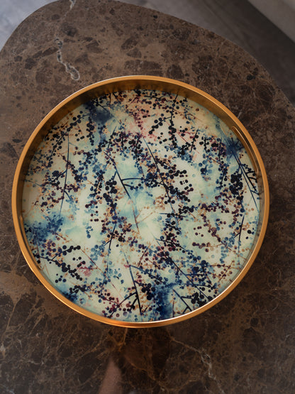 A round decorative tray with a gold-toned rim and a blossom pattern of blue, cream, and black, placed on a dark brown marble surface.