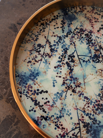 A close-up of a round decorative tray with a gold-toned rim, featuring a printed design of blue berries and blossom on a light cream background with delicate brown branch details.