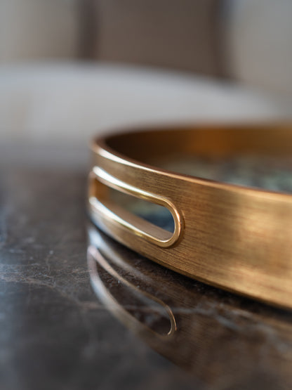 A close-up of a wide, brushed gold tray with cut out handle detail, resting on a dark, reflective marble surface.