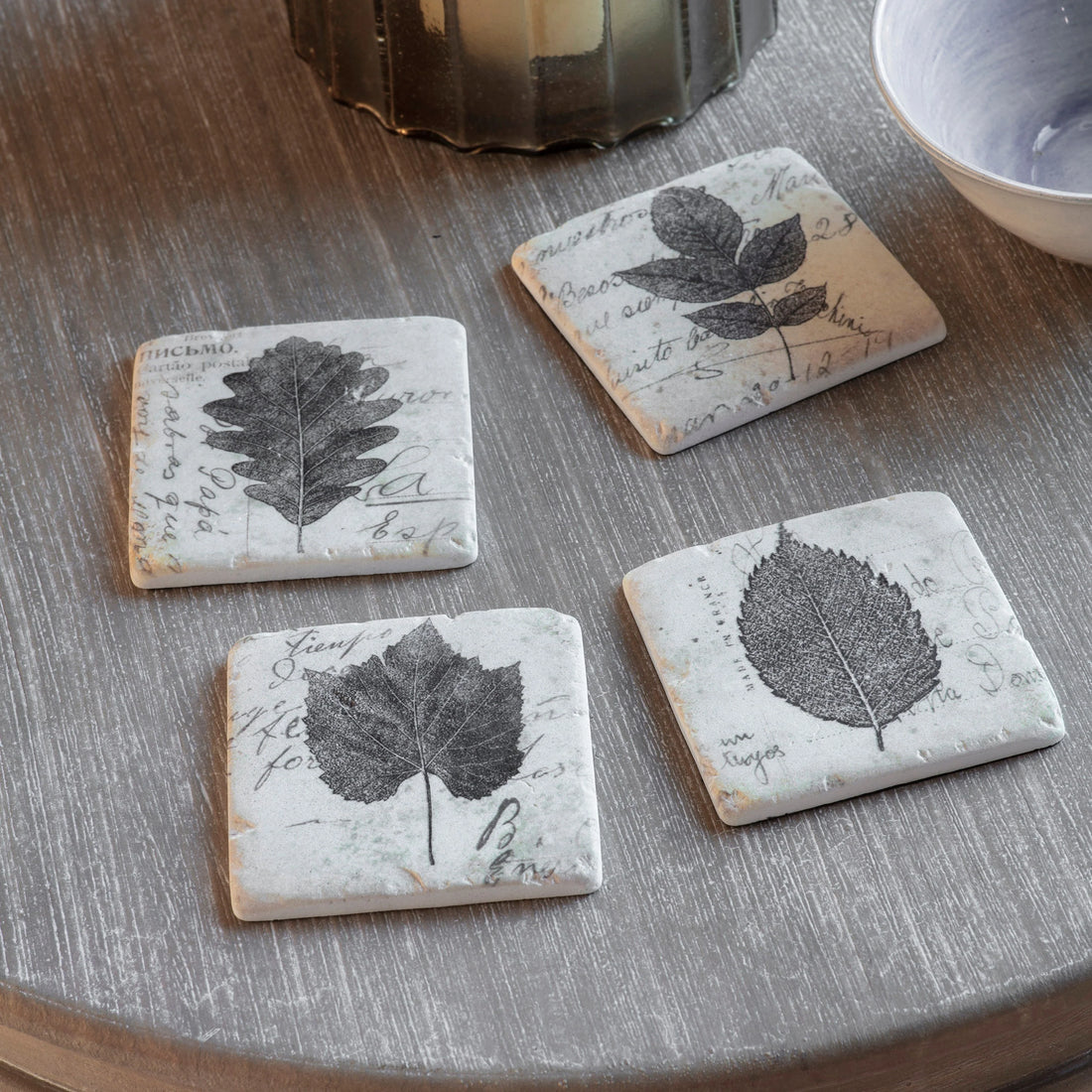 Four leaf-patterned coasters on a wooden surface with a candle and bowl in the background.