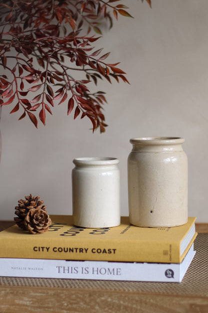 Two cream stoneware jars on a stack of books with a decorative branch and pine cone in the background.