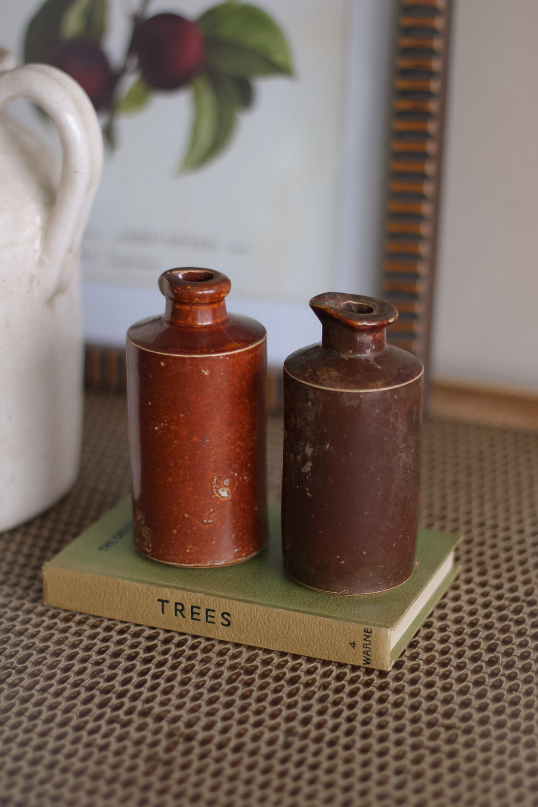 Two brown ceramic bottles on a green book with a white pitcher and fruit painting in the background.