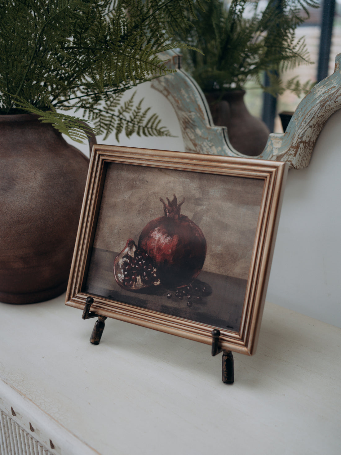 A framed still life print of a pomegranate on a tabletop, styled with potted ferns and a rustic mirror in the background.