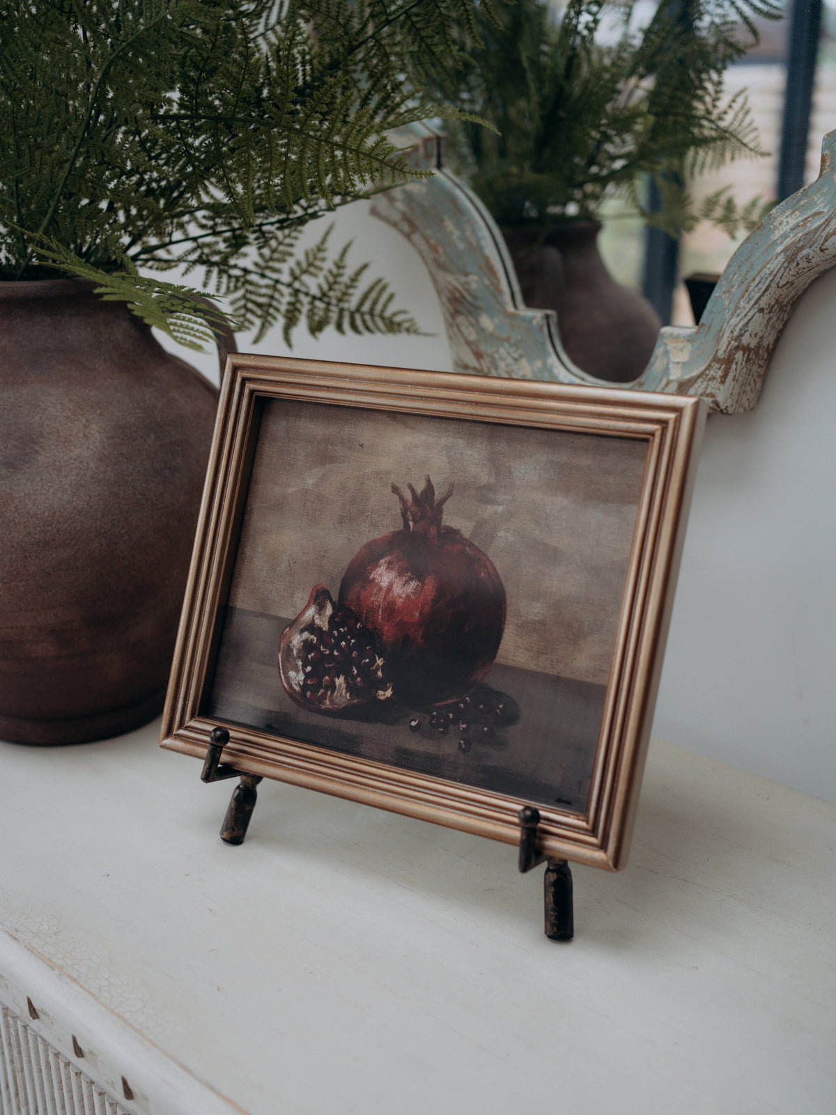 A framed still life print of a pomegranate on a tabletop, styled with potted ferns and a rustic mirror in the background.